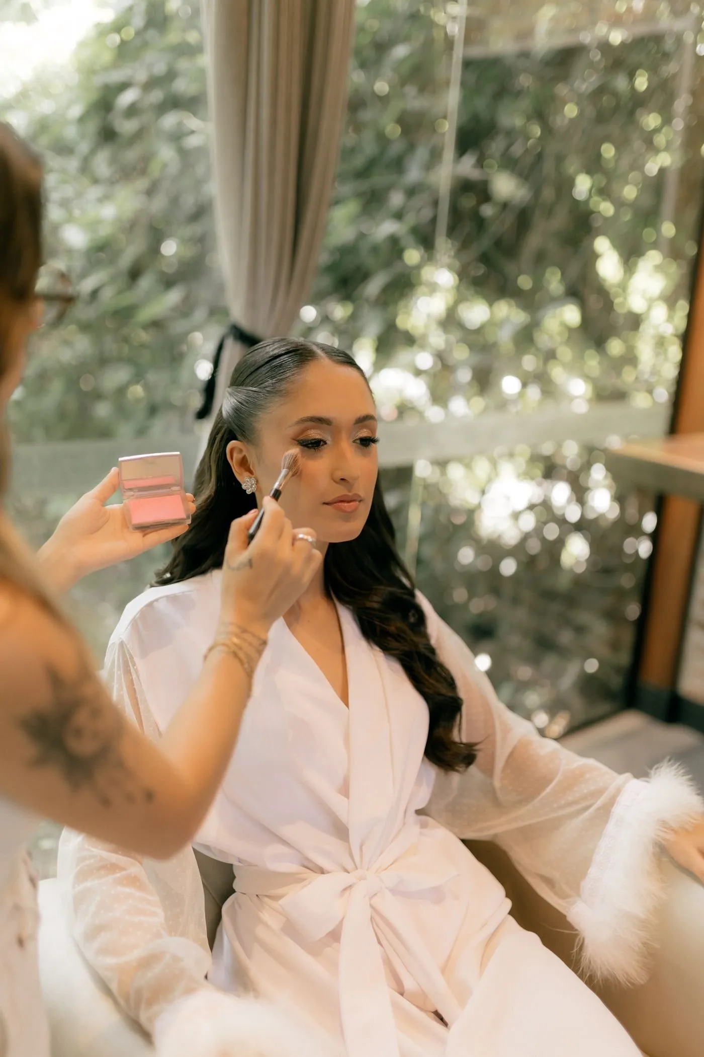 Bride in a white robe having makeup applied beside a sunny window